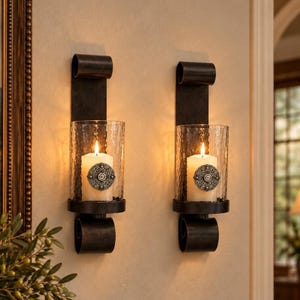 May include: Two dark metal wall sconces featuring a vertical rectangular backplate with curled ends at the top and bottom. Each sconce holds a clear, textured glass cylinder containing a white pillar candle adorned with a round silver medallion.