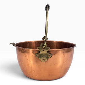 May include: A round copper pot with a polished finish, featuring a brass handle attachment stamped with the word REVERE. The pot includes a tall, arched brass handle with a dark grip at the top and a small pouring spout on the left side.