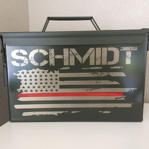 May include: A green metal ammo can with a distressed American flag and red stripe design. The name "SCHMIDT" is printed in silver on the front of the can.