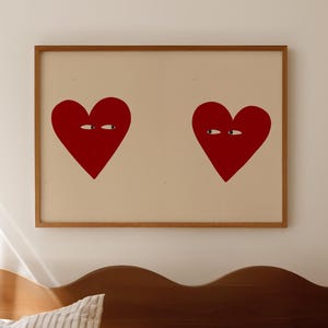 Puede incluir: Una obra de arte enmarcada con dos corazones rojos con ojos sobre un fondo crema. La obra cuelga sobre una cama con ropa de cama a rayas y una cabecera de madera.