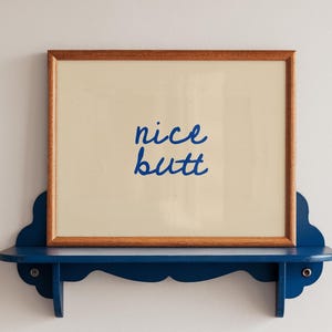 May include: A framed print with the words "nice butt" in blue script. The frame is brown, and the print is displayed on a blue shelf with decorative edges. The shelf is mounted on a white wall.