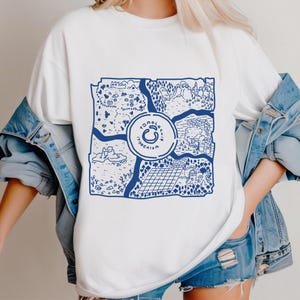 Op de afbeelding: Wit T-shirt met een blauw kaartontwerp en de tekst "Tulum Yucatan" in een cirkelvormig embleem. De kaart toont verschillende landschappen en bezienswaardigheden. Het T-shirt wordt gedragen door een persoon in een spijkerjasje en shorts.