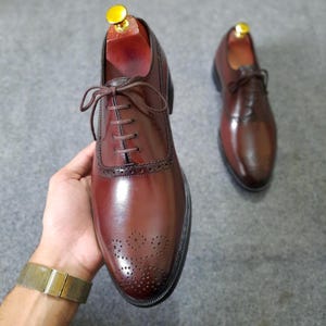 以下が含まれることがあります： バーガンディ色の革製ドレスシューズ。つま先と側面に精巧なブローグのディテールが施されています。靴は茶色の靴紐と黒いソールです。ゴールドカラーのシューツリーが片方の靴に挿入されています。