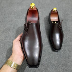 Könnte beinhalten: Ein Paar dunkelbraune Lederschuhe mit poliertem Finish. Die Schuhe haben ein klassisches Design mit eckiger Spitze und Schnürsenkeln. Schuhspanner mit goldfarbenen Knöpfen sind in die Schuhe eingesetzt.