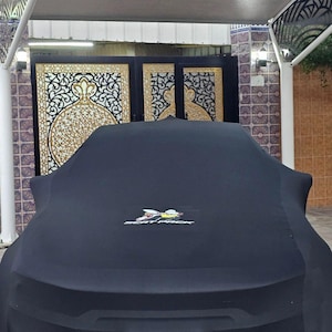 Op de afbeelding: Een zwarte autohoes met een logo van een bij en de tekst "BEAT RICK" bedekt een voertuig. De auto staat onder een witte carport met decoratief metaalwerk. Op de achtergrond zijn sierlijke goud-zwarte poorten te zien.