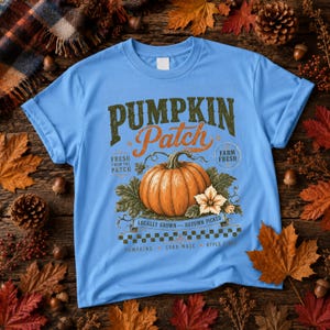 May include: A light blue t-shirt featuring a graphic of an orange pumpkin with green leaves and a white flower. The text reads PUMPKIN Patch, FRESH FROM THE PATCH, FARM FRESH, LOCALLY GROWN, AUTUMN PICKED, PUMPKINS, CORN MAZE, and APPLE CIDER.