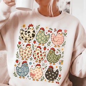 Op de afbeelding: Een beige sweater met ronde hals voorzien van een grafische print van negen kippen in diverse patronen, waaronder koeienvlekken, bloemen, vichy-ruit, kersen, aardbeien, paisley, strikjes en sterren, afgewisseld met kleine bloemetjes, blaadjes en hartjes.