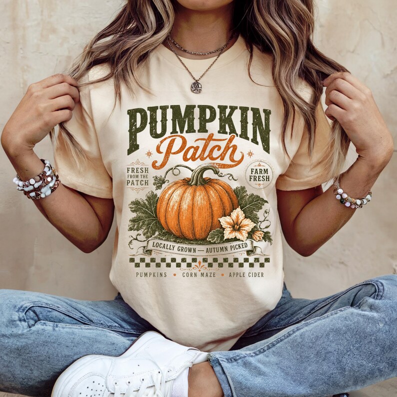 May include: A cream-colored short-sleeve t-shirt featuring a graphic print with the text PUMPKIN Patch, Fresh from the patch, Farm fresh, Locally grown, Autumn picked, Pumpkins, Corn maze, and Apple cider, centered around a large orange pumpkin.