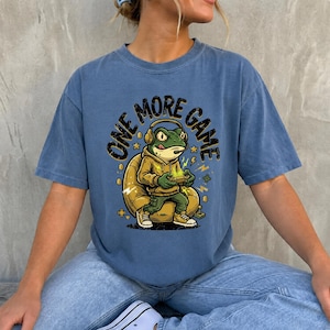 May include: A blue short-sleeve t-shirt featuring a graphic of a green frog wearing a yellow hoodie and headphones while sitting in a chair and playing a video game. The text "ONE MORE GAME" is printed in black above the frog illustration.