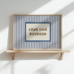 May include: A rectangular framed print featuring a light blue and white vertical striped background. In the center, a cream-colored rectangular label with a jagged blue border displays the brown text "LONG LIVE BOYHOOD" in a bold, rounded font.