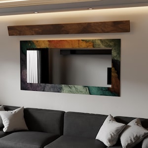 Può includere: Uno specchio rettangolare con una cornice colorata, simile alla pietra, è appeso sopra un divano grigio scuro con cuscini bianchi. Una mensola di legno è montata sopra lo specchio. La cornice dello specchio presenta sfumature di verde, arancione e grigio.