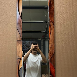 May include: A rectangular mirror with a decorative wooden frame. The frame features a pattern of wood grain and orange accents. The mirror reflects a person holding a phone. The mirror is a vertical design.