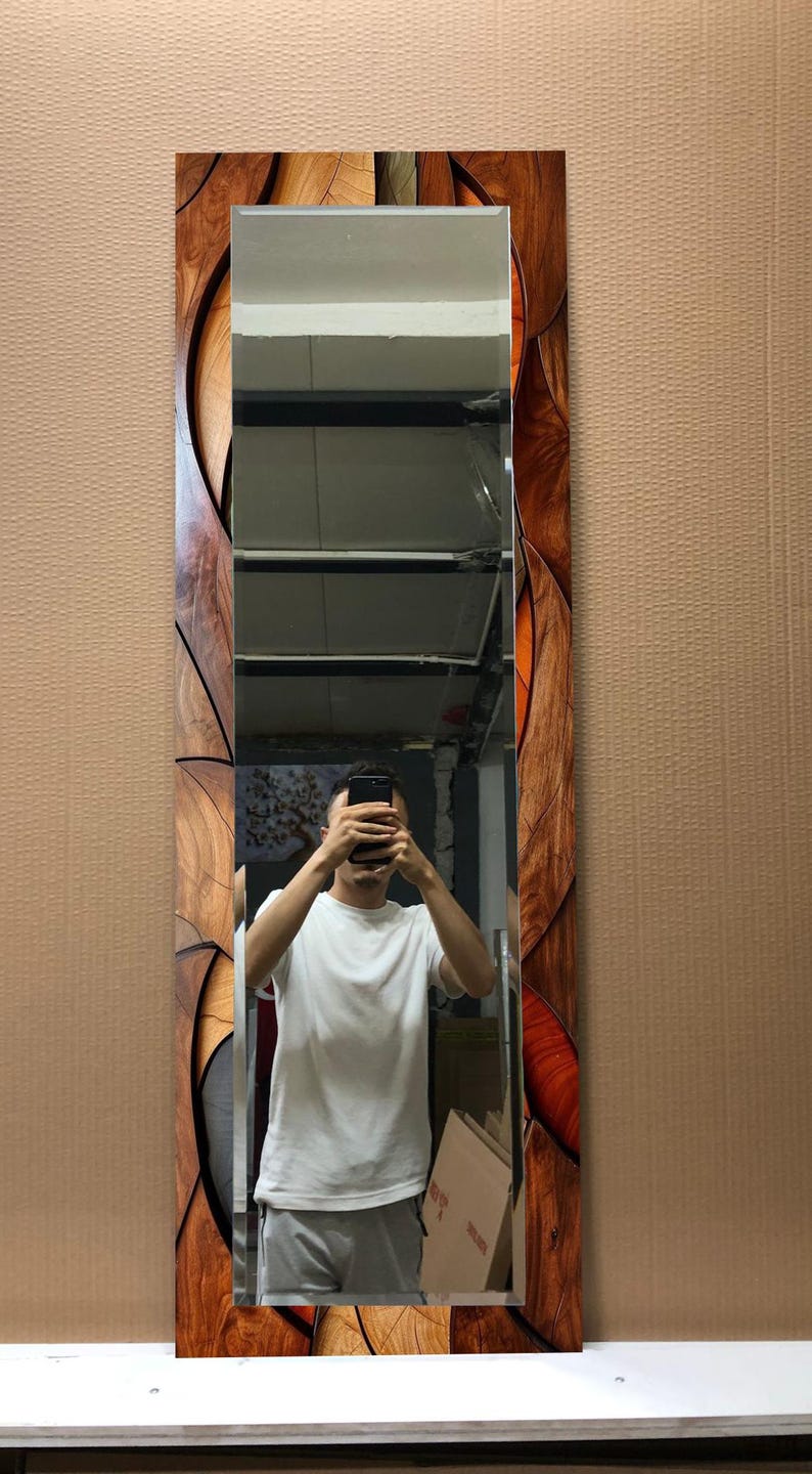 May include: A rectangular mirror with a decorative wooden frame. The frame features a pattern of wood grain and orange accents. The mirror reflects a person holding a phone. The mirror is a vertical design.