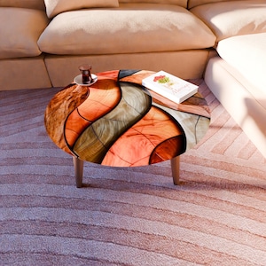 Könnte beinhalten: Ein runder Couchtisch mit einem einzigartigen Holzmaserungsdesign in Orange-, Braun- und Grautönen. Der Tisch hat drei Holzbeine. Ein kleines Buch mit Blumen und eine Tasse Tee stehen auf dem Tisch. Der Durchmesser beträgt ca. 76 cm.