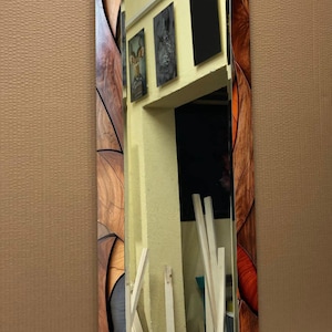 May include: A rectangular mirror with a decorative wooden frame. The frame features a mosaic-like design with various shades of brown and orange. The mirror reflects a room with artwork and white trim.