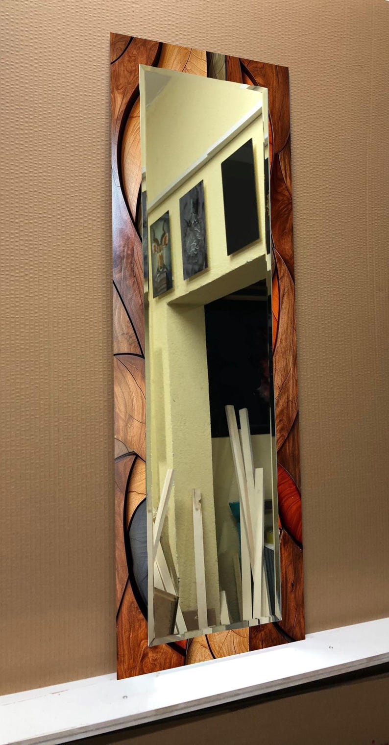 May include: A rectangular mirror with a decorative wooden frame. The frame features a mosaic-like design with various shades of brown and orange. The mirror reflects a room with artwork and white trim.