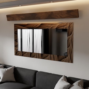 Può includere: Uno specchio rettangolare con una cornice decorativa in legno con un motivo a forma di foglia. Lo specchio è montato su una parete bianca sopra un divano grigio scuro con cuscini grigio chiaro. Un pannello in legno coordinato è sopra lo specchio.