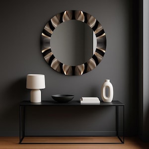 May include: A round decorative mirror with a patterned frame hangs above a black console table. The frame features a repeating design of gold and black shapes. A white lamp, a black bowl, books, and a white vase are on the table.
