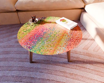 May include: A round coffee table with a colorful, abstract design. The table has a rainbow pattern with shades of red, orange, yellow, green, and pink. It has three wooden legs. A book and a cup of tea sit on the table.