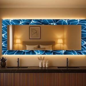 May include: A rectangular mirror with a blue floral design border and a warm, glowing light. The mirror is mounted above a dark countertop with two sinks and a wooden cabinet. The room has a modern, elegant design.