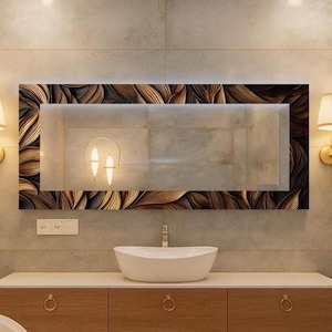 May include: A contemporary bathroom interior with a large rectangular mirror framed with a brown and gold leaf design. The mirror is above a wooden vanity unit with a white oval basin. Gold wall lights with shades are on either side, and a towel rail is present.