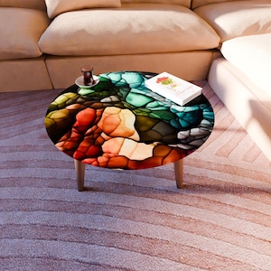 May include: An oval coffee table with a colorful, abstract design. The tabletop features a mosaic-like pattern in shades of red, orange, yellow, green, and blue. The table has light-colored wooden legs. A small book and a cup of tea sit on the table.