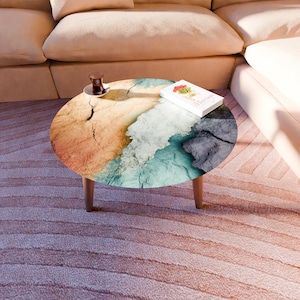 May include: A round coffee table with a unique design. The tabletop features a colorful, textured pattern resembling a coastal landscape with shades of orange, blue, and gray. The table is supported by three wooden legs.