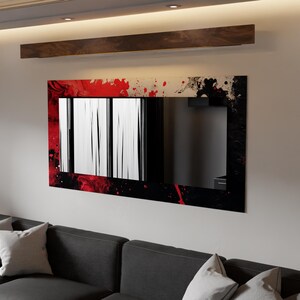 May include: Rectangular wall mirror with a black, red, and white abstract design. The frame features a splatter effect in red and black. The mirror is mounted on a white wall above a dark grey sofa with white cushions.