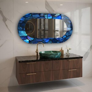 May include: An oval-shaped mirror with a mosaic blue frame hangs above a dark wood vanity. A green vessel sink and gold faucet are visible. The vanity has four drawers with gold handles. The wall is white marble.