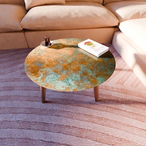 Puede incluir: Una mesa de centro redonda con una superficie de color cobre y turquesa. La mesa tiene tres patas de madera. Un libro con un diseño floral y un vaso pequeño de té están sobre la mesa.