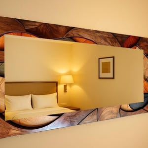 May include: A rectangular wall mirror with a decorative wooden frame. The frame features a mosaic-like pattern in shades of brown, orange, and gray. The mirror reflects a bedroom scene with a bed, lamp, and artwork.