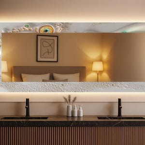 Puede incluir: Espejo rectangular iluminado con un borde blanco texturizado y una sección superior decorativa. El espejo refleja una habitación con una cama, lámparas y obras de arte. El espejo está montado sobre una encimera oscura.
