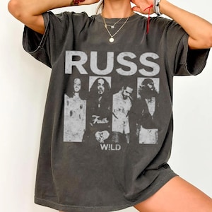 May include: Dark gray t-shirt featuring a band graphic. The design includes the band name "RUSS" in large, distressed white letters above four grayscale images of the band members. Below the images, the words "Faith" and "WILD" are printed.