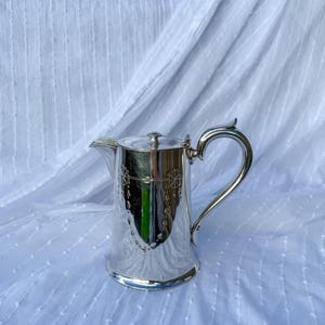 Può includere: Una brocca color argento con un manico curvo e un coperchio. La brocca ha un beccuccio per versare e motivi decorativi incisi. Lo sfondo è un tessuto bianco strutturato.