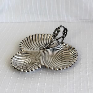 Op de afbeelding: Een zilverkleurige serveerschaal met drie secties en een handvat. De schaal heeft een schelpachtig ontwerp met geribbelde details en een kralenrand. De handgreep heeft sierlijke krullen. De schaal is waarschijnlijk van metaal.