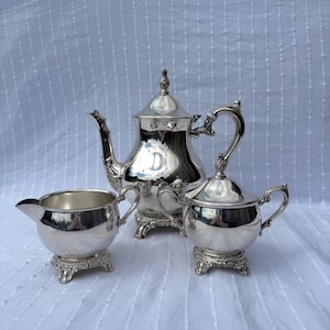 May include: A silver tea set, including a teapot, creamer, and sugar bowl. The set has ornate handles and rests on decorative feet. The reflective surfaces of the set create a bright, elegant appearance. The teapot has a tall, pointed lid.