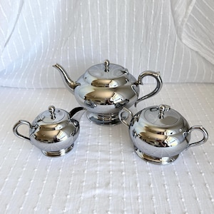 Puede incluir: Un juego de té plateado de tres piezas. El juego incluye una tetera con boquilla y asa, una crema y un azucarero, todos con tapas a juego. Las piezas son muy reflectantes, con un acabado brillante similar a un espejo.