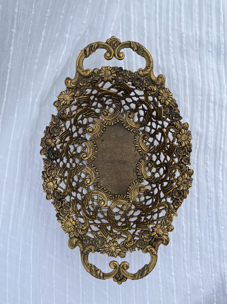 May include: An antique brass oval tray with ornate floral and openwork lace design. The tray features two curved handles and a central rectangular area. The brass has a warm, aged patina, suggesting vintage craftsmanship. The tray is suitable for decorative purposes.