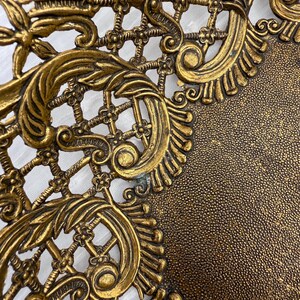 May include: Close-up of a gold-colored, ornate metal decorative piece. The design features intricate scrollwork, floral elements, and a lattice pattern. The surface has a textured appearance, with a darker, speckled area.