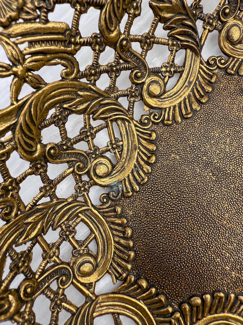 May include: Close-up of a gold-colored, ornate metal decorative piece. The design features intricate scrollwork, floral elements, and a lattice pattern. The surface has a textured appearance, with a darker, speckled area.