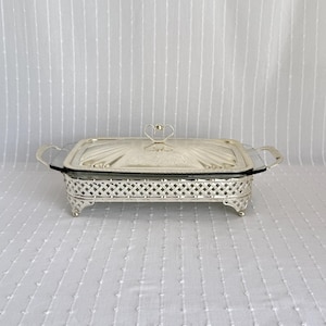 Puede incluir: Una fuente rectangular plateada con un inserto de vidrio y una tapa decorativa. La fuente tiene detalles ornamentados, incluyendo un patrón similar a una celosía alrededor de la base y un asa en forma de corazón en la tapa. La fuente está sobre una superficie blanca.