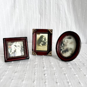 以下が含まれることがあります： 暗い赤色の木製フレームの3つのアンティークフォトフレーム。1つは正方形、1つは長方形、1つは楕円形です。各フレームには、ヴィンテージの白黒写真が収められています。フレームは、白くテクスチャ加工された表面に表示されます。