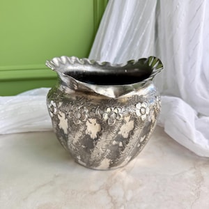 May include: A silver-toned metal bowl featuring a ruffled, scalloped rim and a rounded body. The exterior surface is decorated with embossed floral patterns and textured vertical bands, creating a detailed, metallic finish throughout the piece.