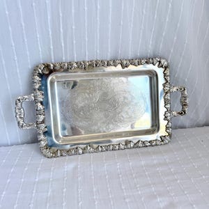 May include: A rectangular silver serving tray with ornate handles and a decorative border. The tray features an embossed floral design in the center. The tray is approximately 12 inches long and 8 inches wide.