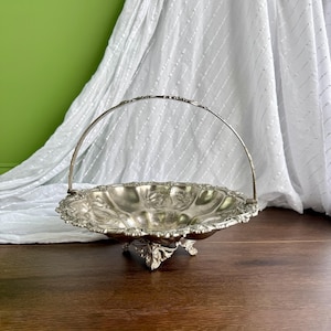 May include: A silver-toned metal basket with a scalloped edge and an arched handle. The bowl features an embossed floral pattern and rests on three ornate, decorative feet. The surface has a polished metallic finish with intricate detailing.
