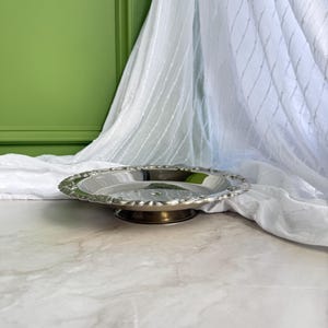 May include: A circular silver-toned metal serving tray with a raised, scalloped edge and a smooth, reflective interior surface. The tray is supported by a short, circular pedestal base, providing a slightly elevated profile for display purposes.