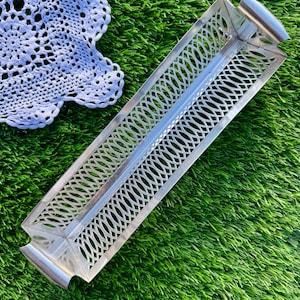 Könnte beinhalten: Ein rechteckiges Tablett aus silberfarbenem Metall mit einem dekorativen durchbrochenen Design aus ovalen Formen. Das Tablett hat gebogene Griffe an jedem Ende. Eine weiße Spitzendecke ist teilweise in der oberen linken Ecke zu sehen, vor dem Hintergrund von grünem Gras.