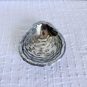 May include: A silver-colored, shell-shaped decorative bowl with a textured surface. The bowl has a hammered appearance and is designed to resemble a clam shell. The bowl is sitting on a white, textured surface.