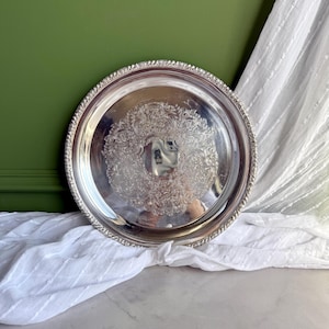 Puede incluir: Una bandeja de servicio redonda de color plateado con un centro pulido y reflectante y un intrincado patrón floral en relieve alrededor del centro. El borde de la bandeja está decorado con un diseño detallado de cuerda.