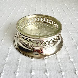 Puede incluir: Un posavasos plateado con un inserto de vidrio. El posavasos tiene un diseño calado decorativo alrededor del borde y una base circular. El inserto de vidrio tiene un patrón de estrella. El posavasos está sobre una superficie blanca texturizada.